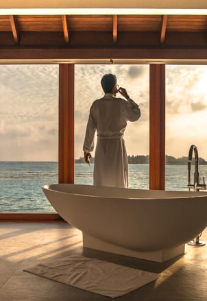 Man in a dressing gown drinking coffer looking at towards sea views from a luxury bathroom.