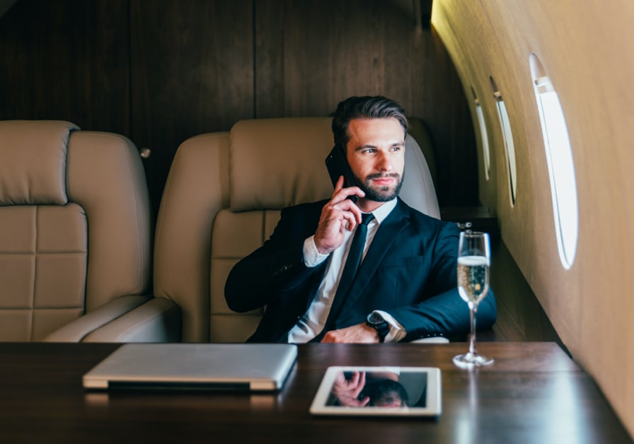 Young, smartly dressing business on the phone on a private jet, with computer and glass of champagne in front of him.